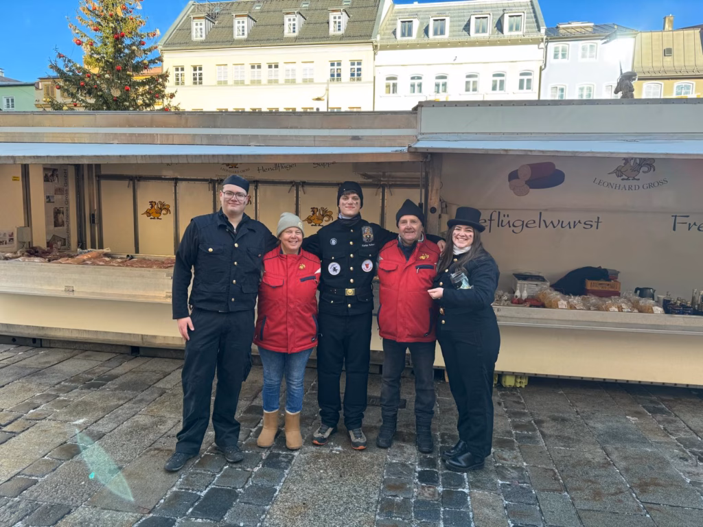F&uuml;nf Personen vor dem Marktstand &bdquo;Leonhard Gro&szlig;&ldquo;, drei davon in Kaminkehrer-Kleidung